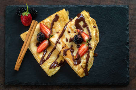 Appetizing thin pancake dessert with hot melted chocolate and red fruit dressing. Top view of crepes on dark tile.の写真素材