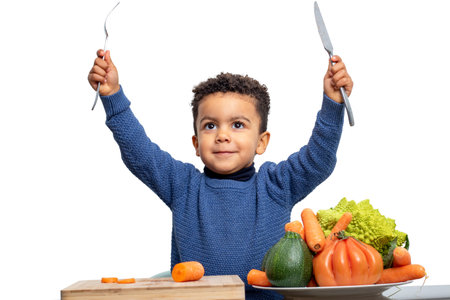 Close up fun portrait of cute afro american kid raising knife and fork with vegetables on table. Boy isolated on white background.の写真素材