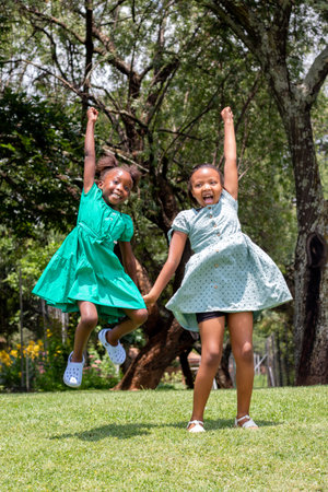 Full-length portrait of two African girls jumping together in a green garden. Two youngsters holding hands shouting.の写真素材