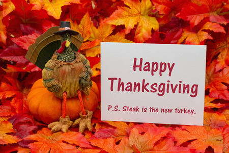 Some fall leaves and a turkey sitting on a pumpkin and a gift tag with a blank gift tag with text Happy Thanksgiving PS Steak is the new turkeyの写真素材