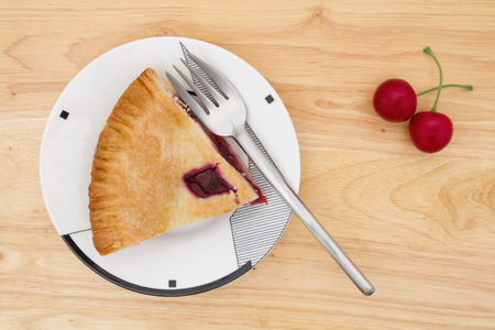 Slice of cherry pie on a plate with a fork and cherries on a wood counter top backgroundの写真素材