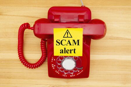 Scam alert message on a sticky note on a red old retro rotary landline phone on a wood deskの写真素材