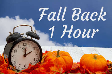 Fall Back 1 hour message with fall leaves, alarm clock, and pumpkins on weathered wood with skyの写真素材