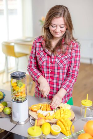 Young smiling woman making smoothie with fresh greens in the blender in kitchen at home.の写真素材