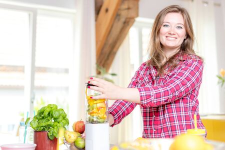 Young smiling woman making smoothie with fresh greens in the blender in kitchen at home.の写真素材