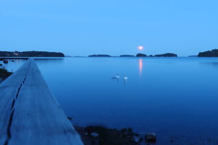 Blue sky and blue water with swans on a lake in Helsinki, Finland. A red sun
and reflection in the middle of the night during summer.の写真素材