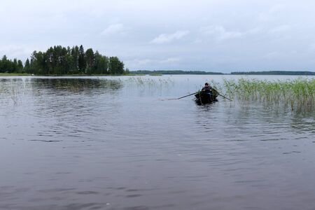 Teenage boy (16) rowing through the weeds on Lake Saimaa, Finland on a cloudy summer day in July with trees in the background.のeditorial素材
