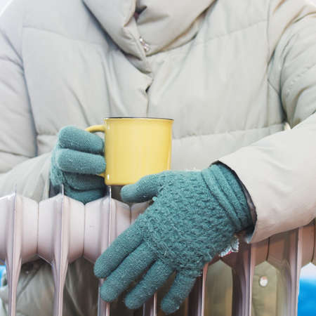 Hands in wool gloves with a cup of tea over a heating radiator. Focus on hands. Concept of heating season.の写真素材