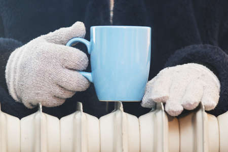 Hands in wool gloves with a cup of tea over a heating radiator. Focus on hands. Concept of heating season.の写真素材
