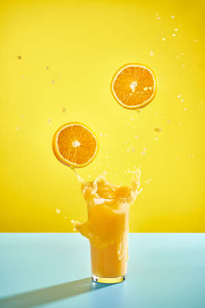 a glass of splashing orange juice and flying oranges on the yellow backgroundの写真素材