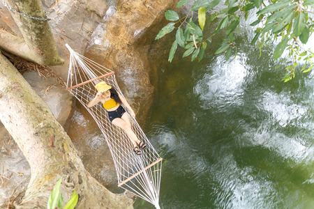 beauty asian woman in yellow knitwear and yellow hat and glasses sitting on cradle,it hanging on tree near streamの写真素材