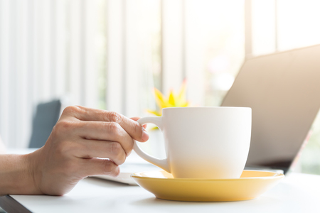worker holding cup of coffee after break and laptop on desk near window in cafeの写真素材
