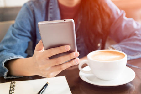 close up hand of business asian woman use smartphone and white cup on wooden table in coffee shop with light effect and vintage toneの写真素材