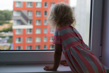 Small curly girl with blond hair looking outside from the window in multistory neighbourhoodの写真素材