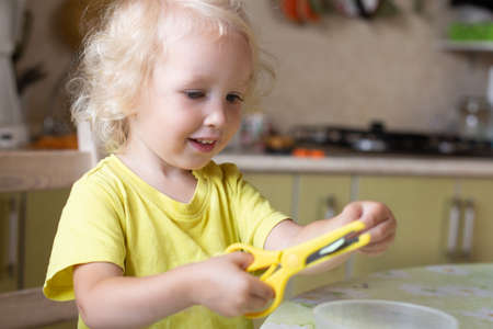 Toddler girl with blond curly hear, wearing yellow t-shirt, learning how to use scissorsの写真素材