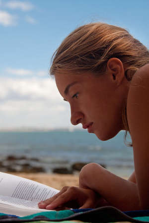 Portrait of young woman thoroughly reading book laying on the beach. Sea with stones is on the...