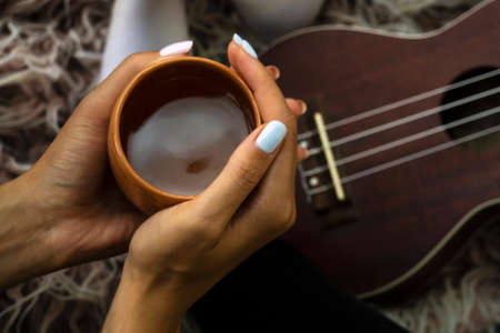 Cozy photo of womans hands holding cup of tea with ukulele on backdrop. Fall or winter time concept.