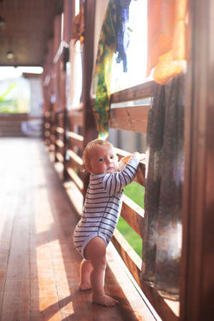 Baby boy making his first steps in sunny morning. Boy is standing barefoot outside in the country house in the sunlight. Drying cloths are on the background. Cozy warm mood, vertical formatの写真素材