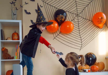 Boy in a black cape and a witchs hat glues a paper bat to the wall. The girl helps her brother, gives him a bat.の写真素材