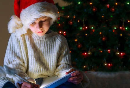 Cute boy in red santa hat and white sweater writing letter to Santa on the background of a Christmas tree decorated with lights.の写真素材