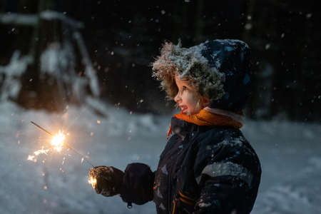 Toddler girl in winter clothes walking outside and holding sparkler in her hand. It is dark and snowy, girl is happily smiling. Magic winter holidays mood