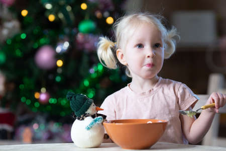 Cute blond toddler girl with blue eyes having her meal in new year entourage. Christmas tree is standing behind. Girl is left-handed.の写真素材