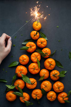 Christmas tree made from tangerines and sparkler, kept by female hand. Flat lay on dark background....