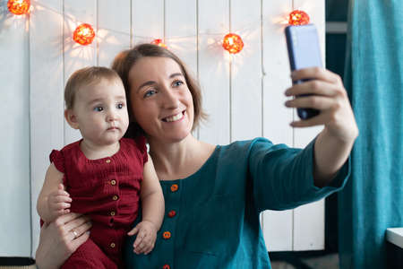 Cheerful woman and her small daughter making selfie in christmas decoration. Winter holidays remote celebration, stay home concept. Mother blogger creating stories. Smartphone and new year backgroundの写真素材