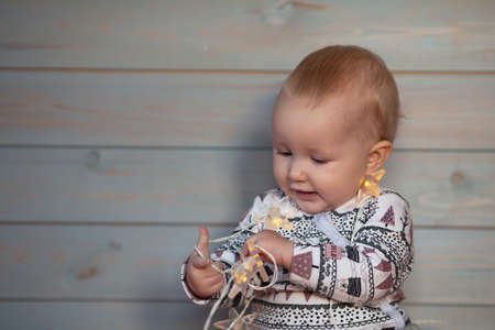 Baby girl wearing clothes with Christmas print playing with lights on light wooden background. New...