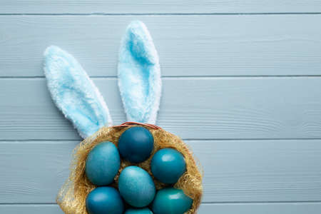 Easter flat lay with blue eggs in the basket and funny bunny ears on light blue wooden background....