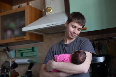 Young man carefully keeping his newborn daughter. Man is standing in the kitchen and smiling. Lifestyleの写真素材