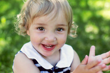 Portrait of cute curly happy toddler girl in sunny summer day against green grass. Baby girl is...
