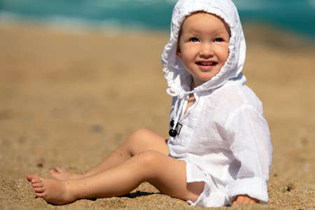 Happy carefree toddler girl in white hoody sitting on the sandy beach. Summer mood. Sea coast. Copy spaceの写真素材