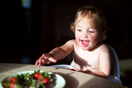 Cute baby girl eating strawberries indoors in the sunlight. Cottagecore aesthetics concept, carefree childhoodの写真素材