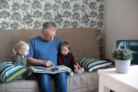 Senior man reading book to small girls. Grandfather spending time with granddaughters on a couch....