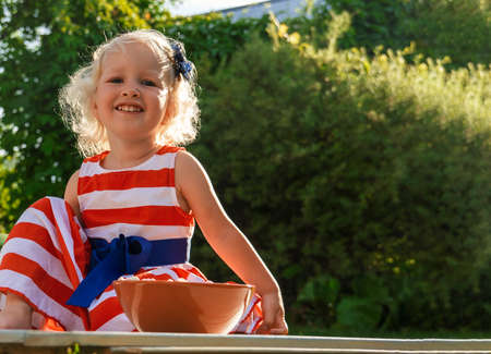 Pretty curly little girl seating in garden with plate of food and looking at the camera.の写真素材