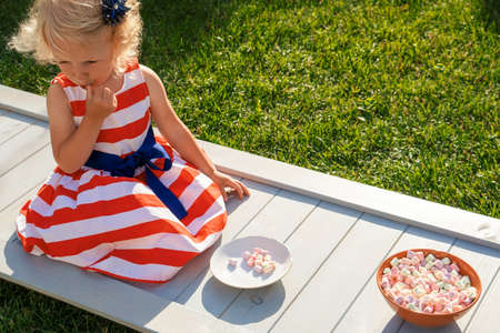 Pretty curly little girl seating in garden and eating marshmallows from the plate.の写真素材
