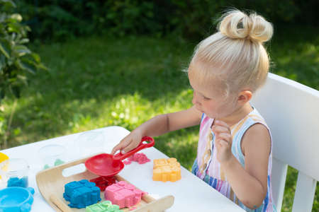 Little cute girl with blonde hair playing with colorful kinetic sand outdoors in the backyard in summer day. Hobby, leisure activity, crafting, modeling and creative hobby conceptの写真素材