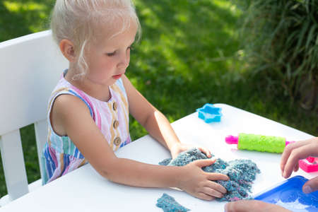 Little cute girl with blonde hair playing with colorful kinetic sand outdoors in the backyard in summer day. Hobby, leisure activity, crafting, modeling and creative hobby conceptの写真素材