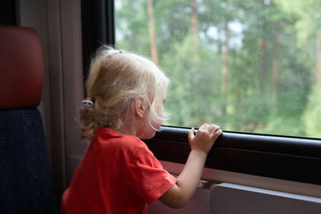Little girl with blonde hair traveling by train. Girl is looking through window. Public...