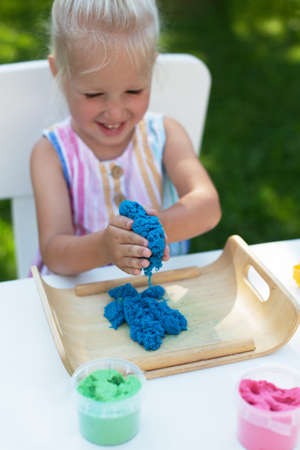 Little cute girl with blonde hair playing with colorful kinetic sand outdoors in the backyard in summer day. Hobby, leisure activity, crafting, modeling and creative hobby conceptの写真素材