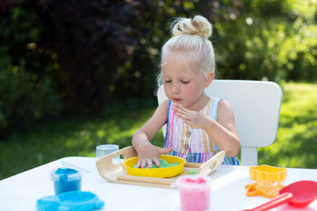 Little cute girl with blonde hair playing with colorful kinetic sand outdoors in the backyard in summer day. Hobby, leisure activity, crafting, modeling and creative hobby conceptの写真素材