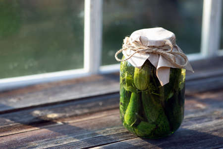 Jar of salted cucumbers standing on old wooden table against terrace window. Harvest season and homemade preserves. Copy space. Pickled vegetablesの写真素材