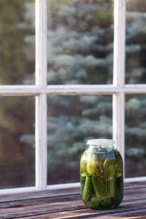 Jar of salted cucumbers standing on old wooden table against terrace window. Harvest season and homemade preserves. Vertical format, copy space. Pickled vegetablesの写真素材