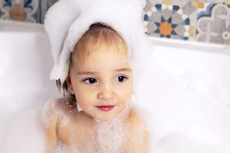 Cute kid with big beautiful eyes sitting in bathroom with bath foam. Baby care, personal care. Girlの写真素材