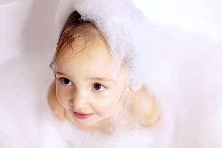 Cute kid with big beautiful eyes sitting in bathroom with bath foam. Baby care, personal care. Girlの写真素材