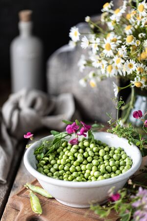 Young green peas in white bowl on wooden background. Pea flowers and daisy flowers on the table.の写真素材