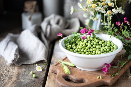 Young green peas in white bowl on wooden background. Pea flowers and daisy flowers on the table.の写真素材