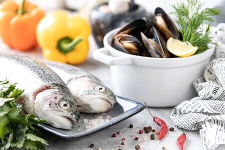 Two raw rainbow trout fish on a plate, greens and fresh vegetables for preparing healthy and tasty food. White pan with mussels on a concrete background. Healthy diet and delicious culinary concept.の写真素材