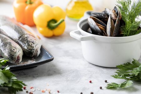 Two raw rainbow trout fish on a plate, greens and fresh vegetables for preparing healthy and tasty food. White pan with mussels on a concrete background. Healthy diet and delicious culinary concept. Copy space.の写真素材